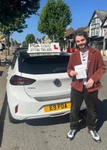 student who passed his driving test with us standing opposite the test centre on a bright sunny day holding his pass certificate and looking excited